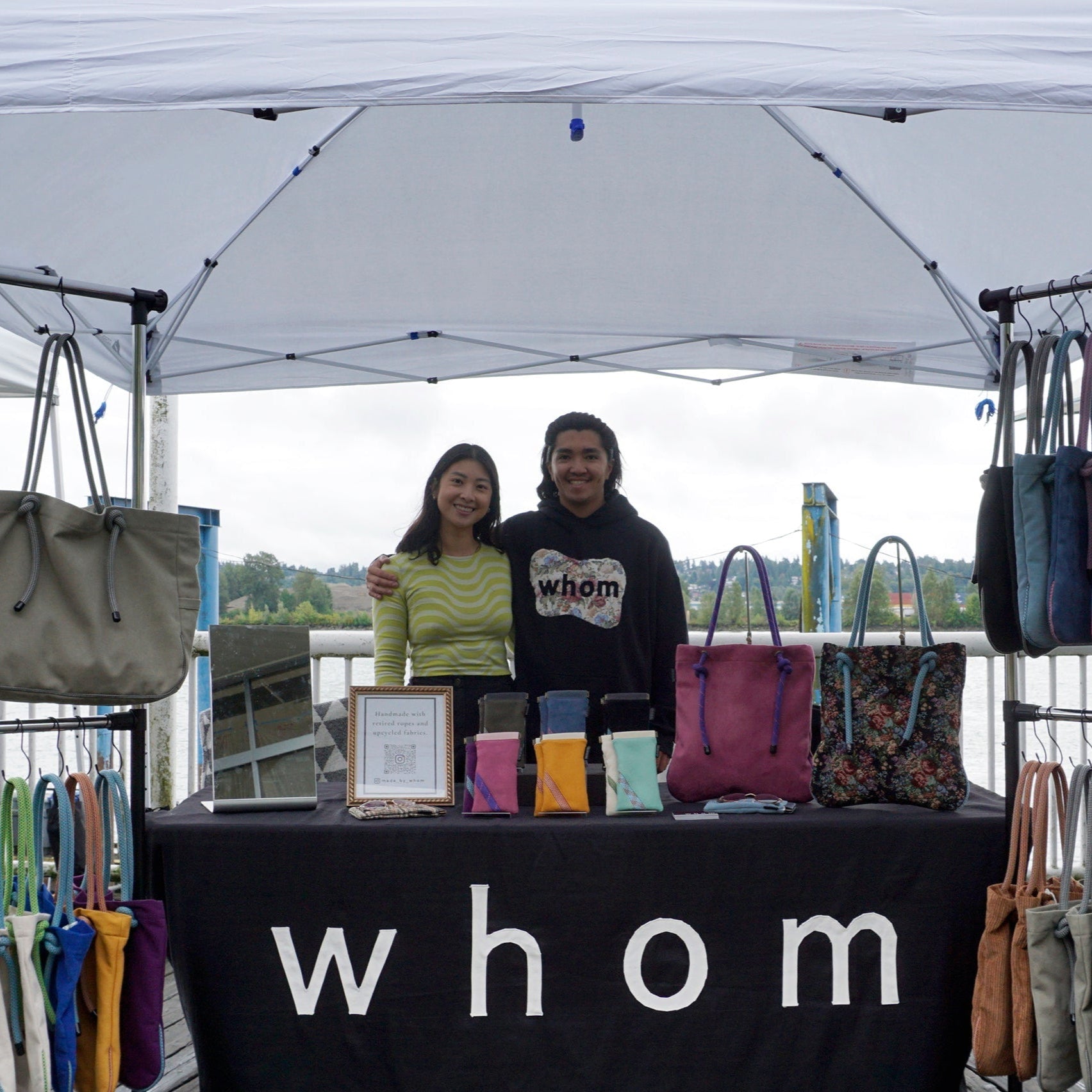Two people standing behind a table with 'whom' branding, surrounded by handbags under a tent.